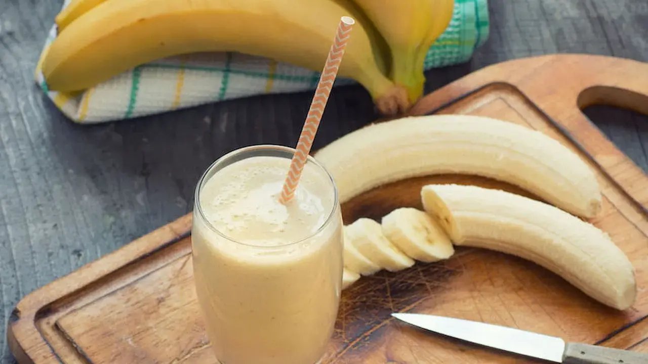 Frullato di banana in un bicchiere con una cannuccia, banane intere e affettate su un tagliere. Con la diarrea si può mangiare la banana, è un alimento delicato.