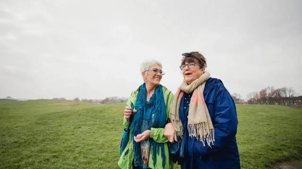Due anziane passeggiano in un prato. Una delle due, con un foulard blu, sembra avere difficoltà a camminare, ma l'altra le offre supporto, un gesto di cura che va oltre l'Alzheimer.