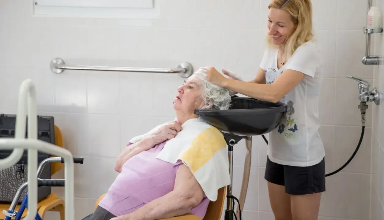 Assistente che lava i capelli a un'anziana non autosufficiente in un bagno attrezzato, con cura e attenzione.