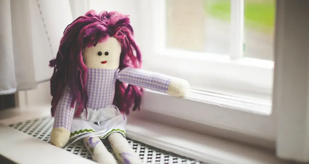 Bambola con capelli viola, vestita a quadretti, seduta sul davanzale. Un simbolo di conforto nella doll therapy per l'Alzheimer.