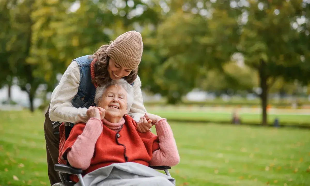Una giovane donna sorride mentre abbraccia un'anziana in sedia a rotelle in un parco. Un momento di gioia che mostra come un caregiver può sopravvivere con amore.