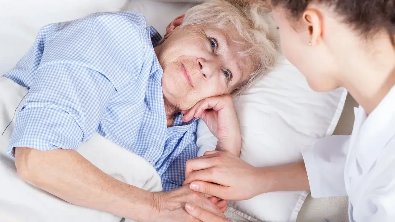 Anziana a letto, assistita da un infermiere. La sua mano stringe quella dell'assistente, forse per alleviare il dolore di una piaga da decubito sull'osso sacro.