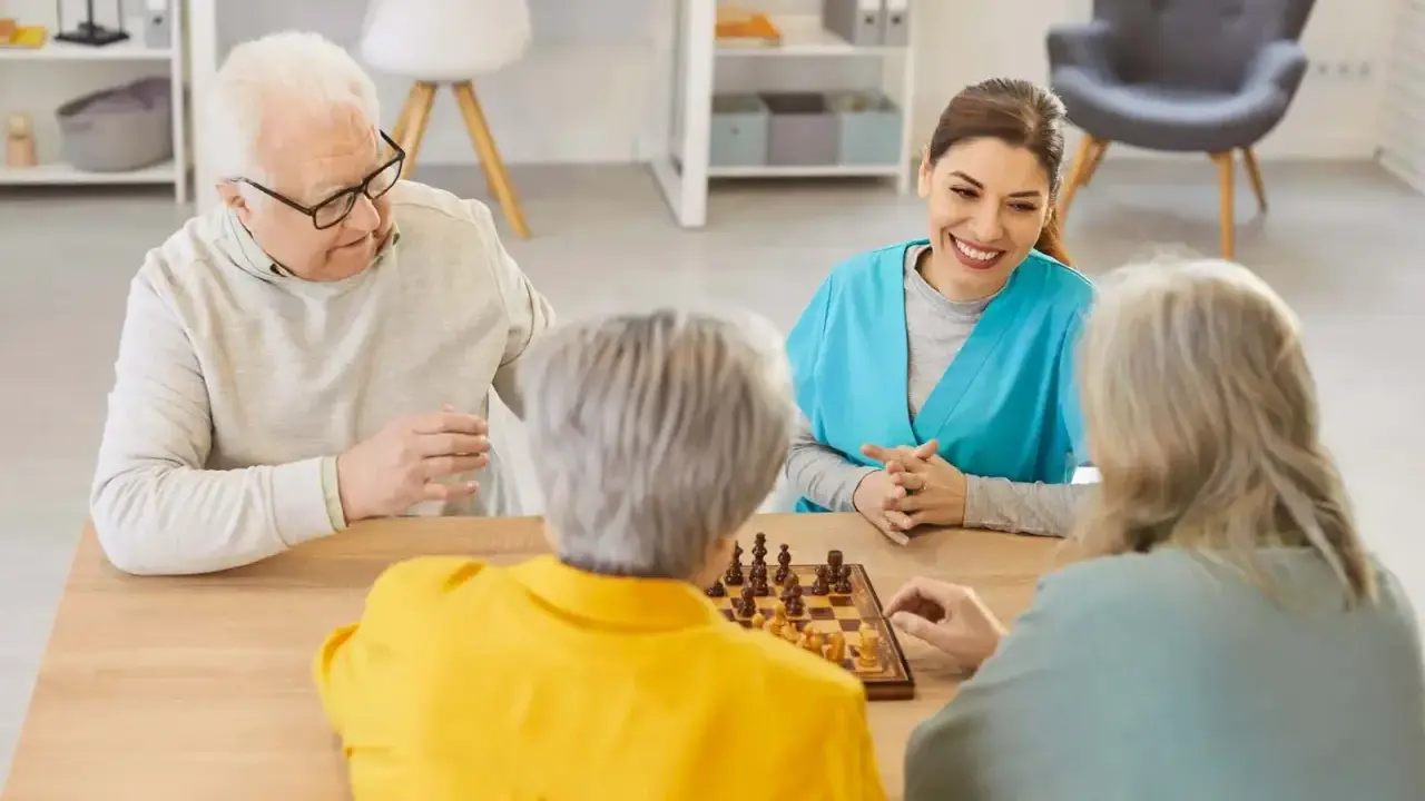 Un gruppo di anziani gioca a scacchi, un'attività che promuove la stimolazione cognitiva. Un'assistente sorride osservando.