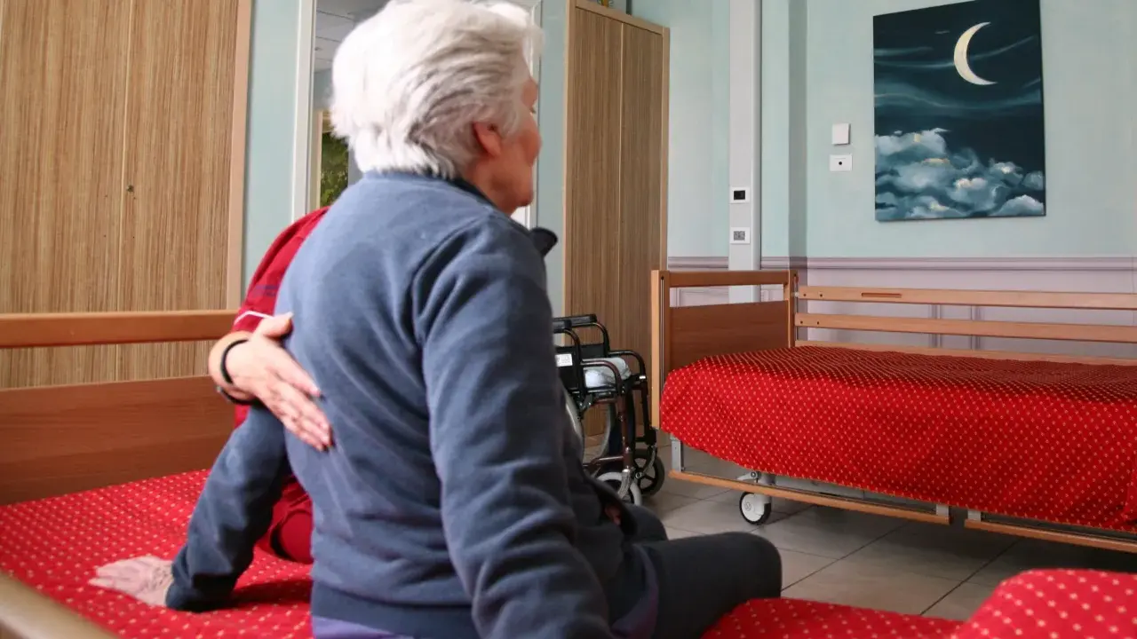 Anziana in stanza, forse affetta da sindrome da ospedalizzazione, con un'altra persona che le appoggia una mano sulla spalla. Sulla parete un quadro con luna e nuvole.