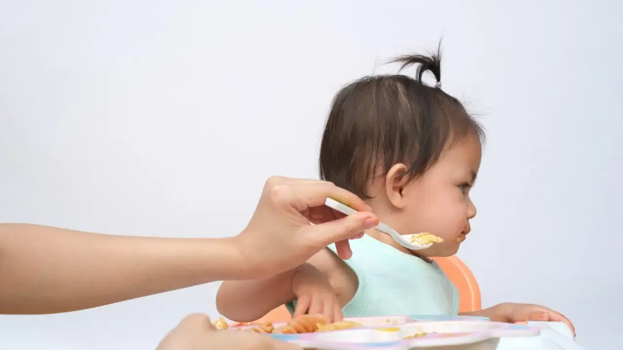 Bambina seduta sul seggiolone, con un cucchiaino di cibo offerto da una mano. Un modo per stimolare l'appetito con pazienza e amore.