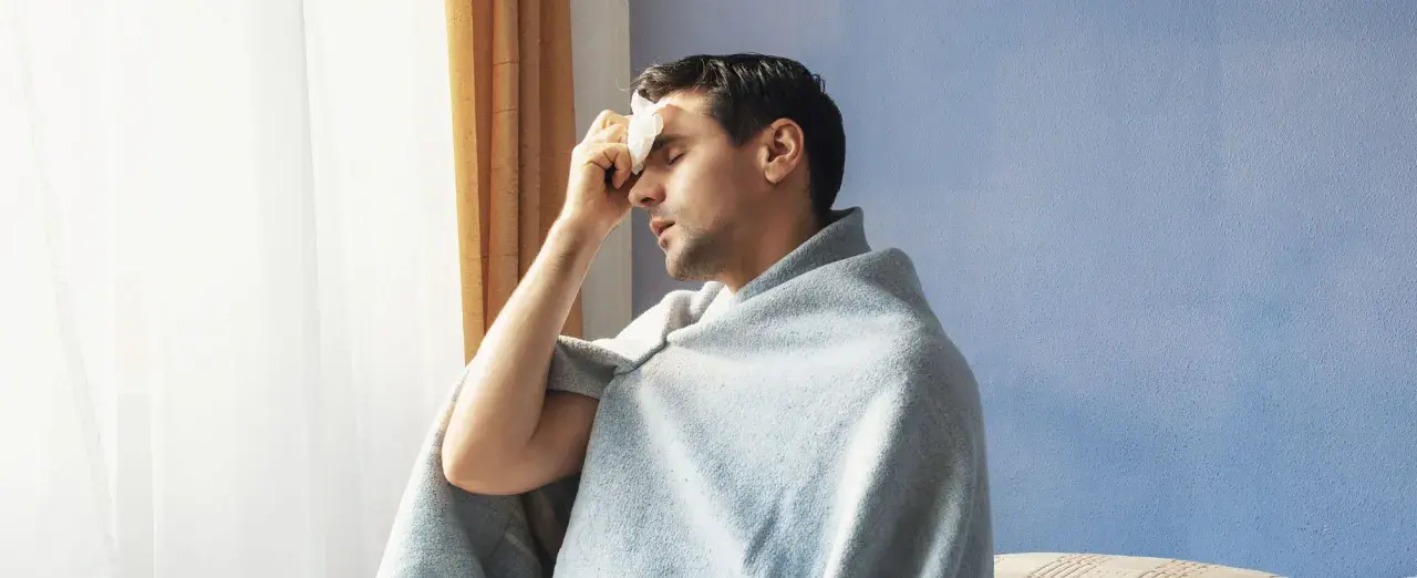 Uomo con panno umido sulla fronte, avvolto in una coperta, che sembra soffrire di sudori freddi.