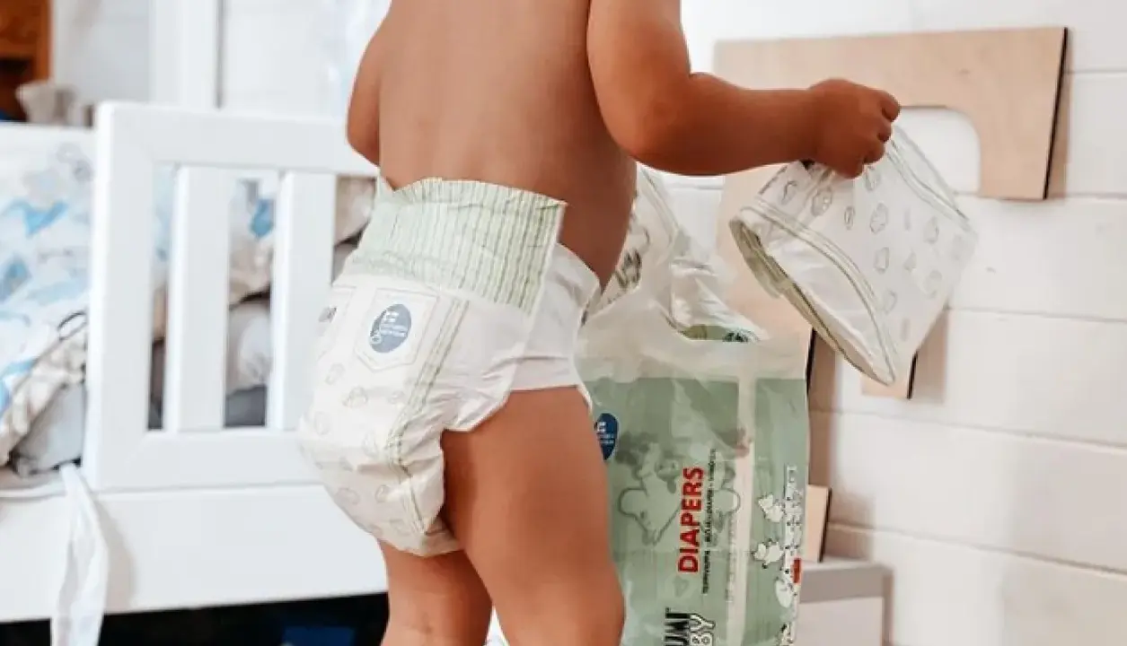 Un bimbo con un pannolino indosso, vicino a una confezione di pannolini. Sembra che i pannolini scadano, ma non c'è fretta.