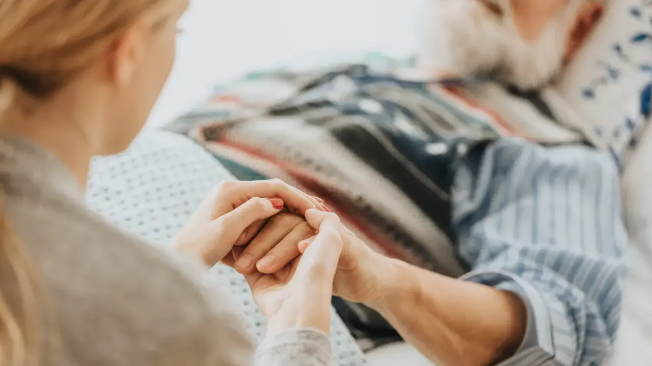 Mani che si stringono tra una giovane donna e un anziano a letto, con cannule nasali. Il caregiver viene pagato per la sua dedizione.