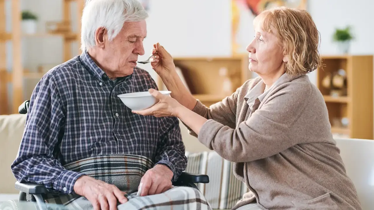 Una donna nutre con un cucchiaino il suo anziano nonno, che non mangia più, seduto su una sedia a rotelle.