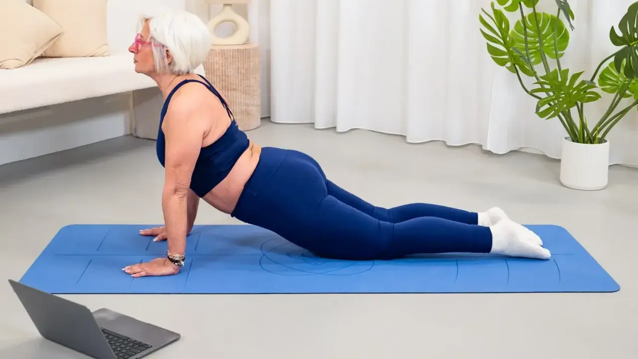 Donna anziana in posa yoga cobra su tappetino blu, seguendo un tutorial online. Un esempio di palestra per anziani attiva e moderna.