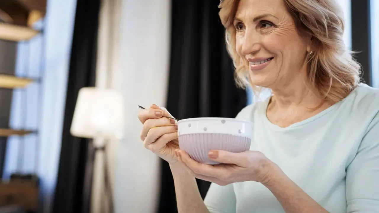 Donna sorride mentre mangia una zuppa cremosa, un esempio di cosa mangiare con disfagia.