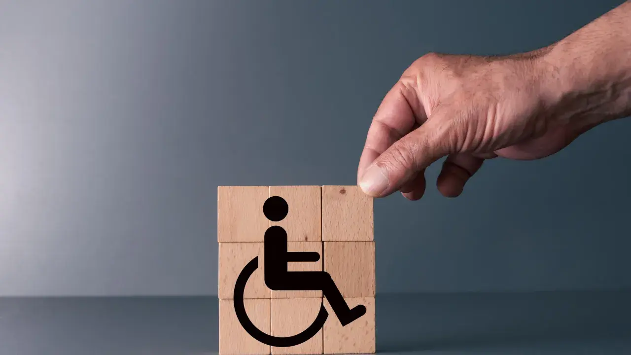 Simboli di accessibilità su cubi di legno, con una mano che aggiunge l'ultimo pezzo. Rappresenta il **certificato medico introduttivo per accompagnamento**.