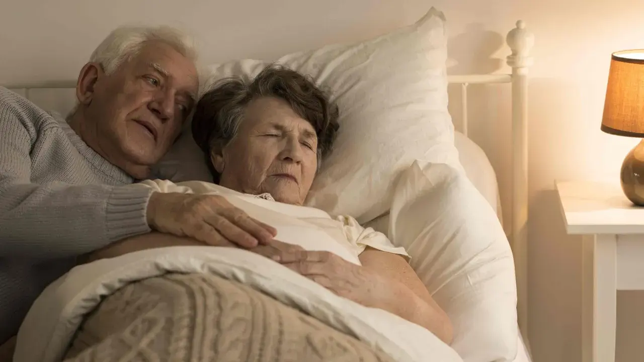 Un uomo anziano abbraccia dolcemente la moglie a letto. La sua mano sulla sua pancia, forse un gesto per confortarla, perché un malato di Alzheimer dorme sempre, cercando riposo.
