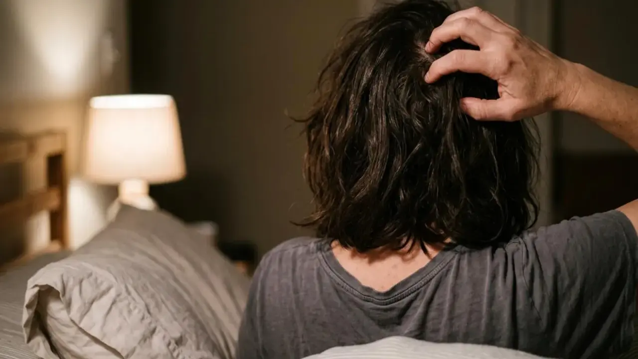 Persona a letto che si gratta la testa, forse perché il prurito aumenta di sera. Luce soffusa da una lampada.