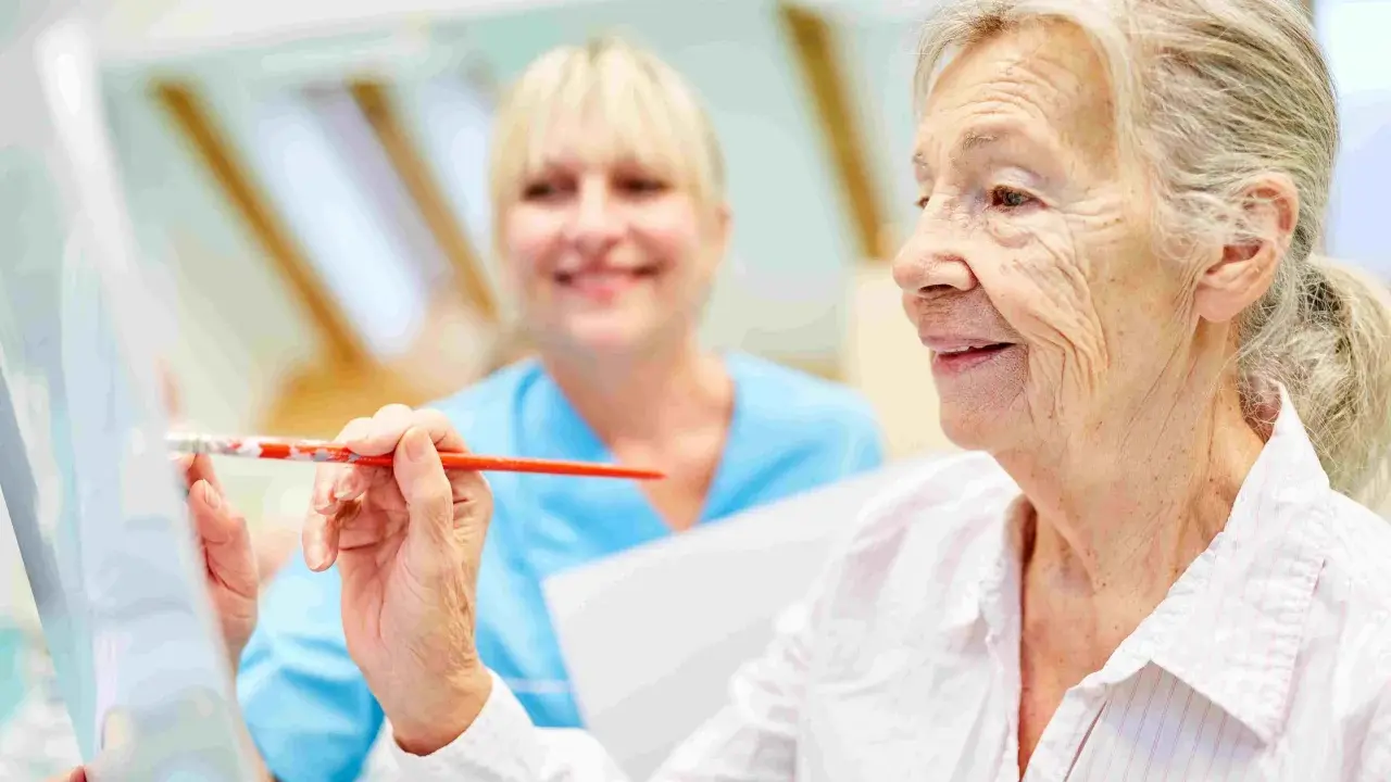 Anziana dipinge con un pennello, assistita da un'infermiera sorridente. Un esempio di terapie non farmacologiche per l'Alzheimer che stimolano la creatività.