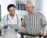 Medico spiega a paziente anziano con deambulatore il significato di un problema cognitivo, mostrando un documento.