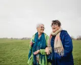 Due anziane passeggiano in un prato. Una delle due, con un foulard blu, sembra avere difficoltà a camminare, ma l'altra le offre supporto, un gesto di cura che va oltre l'Alzheimer.