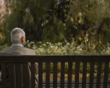 Un anziano seduto su una panchina in un parco, con lo sguardo perso nel vuoto, forse riflettendo sul significato del suo vagare nella vita.