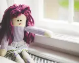 Bambola con capelli viola, vestita a quadretti, seduta sul davanzale. Un simbolo di conforto nella doll therapy per l'Alzheimer.