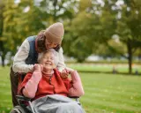 Una giovane donna sorride mentre abbraccia un'anziana in sedia a rotelle in un parco. Un momento di gioia che mostra come un caregiver può sopravvivere con amore.