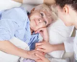 Anziana a letto, assistita da un infermiere. La sua mano stringe quella dell'assistente, forse per alleviare il dolore di una piaga da decubito sull'osso sacro.