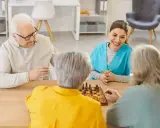 Un gruppo di anziani gioca a scacchi, un'attività che promuove la stimolazione cognitiva. Un'assistente sorride osservando.