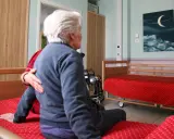 Anziana in stanza, forse affetta da sindrome da ospedalizzazione, con un'altra persona che le appoggia una mano sulla spalla. Sulla parete un quadro con luna e nuvole.