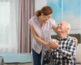 Una donna sorride a un anziano con demenza, tenendo una tazza. Un gesto di cura e affetto.