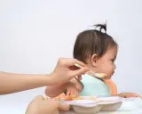 Bambina seduta sul seggiolone, con un cucchiaino di cibo offerto da una mano. Un modo per stimolare l'appetito con pazienza e amore.