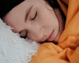 Viso di donna con gocce di sudore sulla fronte, avvolta in una coperta arancione e piumino bianco. Sembra che stia per sudare freddo.