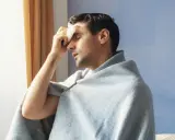 Uomo con panno umido sulla fronte, avvolto in una coperta, che sembra soffrire di sudori freddi.