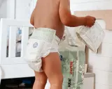 Un bimbo con un pannolino indosso, vicino a una confezione di pannolini. Sembra che i pannolini scadano, ma non c'è fretta.