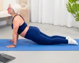 Donna anziana in posa yoga cobra su tappetino blu, seguendo un tutorial online. Un esempio di palestra per anziani attiva e moderna.