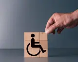 Simboli di accessibilità su cubi di legno, con una mano che aggiunge l'ultimo pezzo. Rappresenta il **certificato medico introduttivo per accompagnamento**.