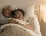 Un uomo anziano abbraccia dolcemente la moglie a letto. La sua mano sulla sua pancia, forse un gesto per confortarla, perché un malato di Alzheimer dorme sempre, cercando riposo.
