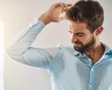Uomo con camicia azzurra macchiata di sudore sotto le ascelle, che si gratta la testa. Sudare tanto è sintomo di stress o caldo.