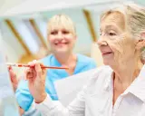 Anziana dipinge con un pennello, assistita da un'infermiera sorridente. Un esempio di terapie non farmacologiche per l'Alzheimer che stimolano la creatività.