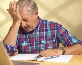 Uomo anziano con mano sulla fronte, frustrato dal non ricordare le cose appena fatte, con quaderno e penna.