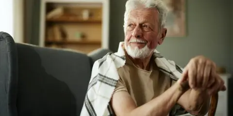 Anziano con bastone, forse per uno dei 10 sintomi parkinson. Sguardo pensieroso, coperto da una coperta a quadri.