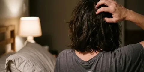 Persona a letto che si gratta la testa, forse perché il prurito aumenta di sera. Luce soffusa da una lampada.