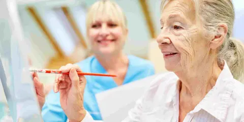 Anziana dipinge con un pennello, assistita da un'infermiera sorridente. Un esempio di terapie non farmacologiche per l'Alzheimer che stimolano la creatività.