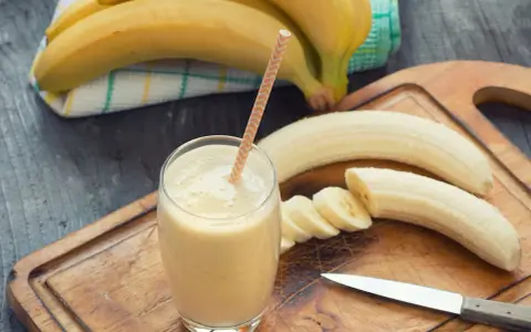 Frullato di banana in un bicchiere con una cannuccia, banane intere e affettate su un tagliere. Con la diarrea si può mangiare la banana, è un alimento delicato.