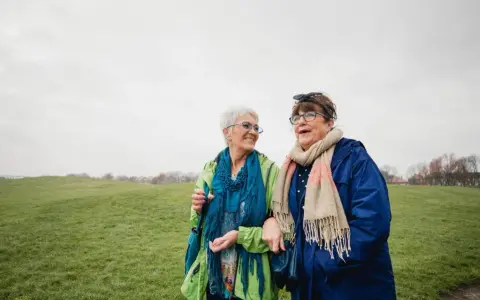 Due anziane passeggiano in un prato. Una delle due, con un foulard blu, sembra avere difficoltà a camminare, ma l'altra le offre supporto, un gesto di cura che va oltre l'Alzheimer.