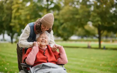 Una giovane donna sorride mentre abbraccia un'anziana in sedia a rotelle in un parco. Un momento di gioia che mostra come un caregiver può sopravvivere con amore.