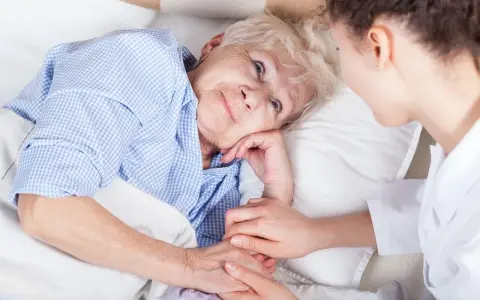 Anziana a letto, assistita da un infermiere. La sua mano stringe quella dell'assistente, forse per alleviare il dolore di una piaga da decubito sull'osso sacro.