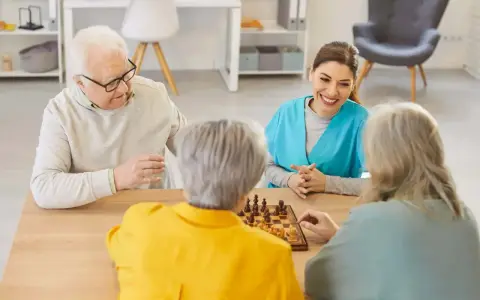Un gruppo di anziani gioca a scacchi, un'attività che promuove la stimolazione cognitiva. Un'assistente sorride osservando.