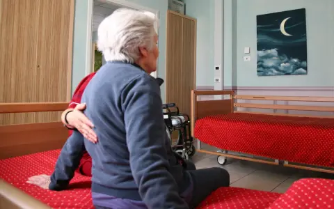Anziana in stanza, forse affetta da sindrome da ospedalizzazione, con un'altra persona che le appoggia una mano sulla spalla. Sulla parete un quadro con luna e nuvole.