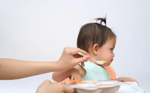Bambina seduta sul seggiolone, con un cucchiaino di cibo offerto da una mano. Un modo per stimolare l'appetito con pazienza e amore.
