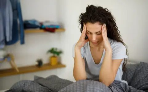 Donna con le mani sulle tempie, seduta a letto, soffre di giramenti di testa.
