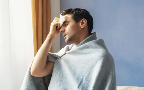 Uomo con panno umido sulla fronte, avvolto in una coperta, che sembra soffrire di sudori freddi.